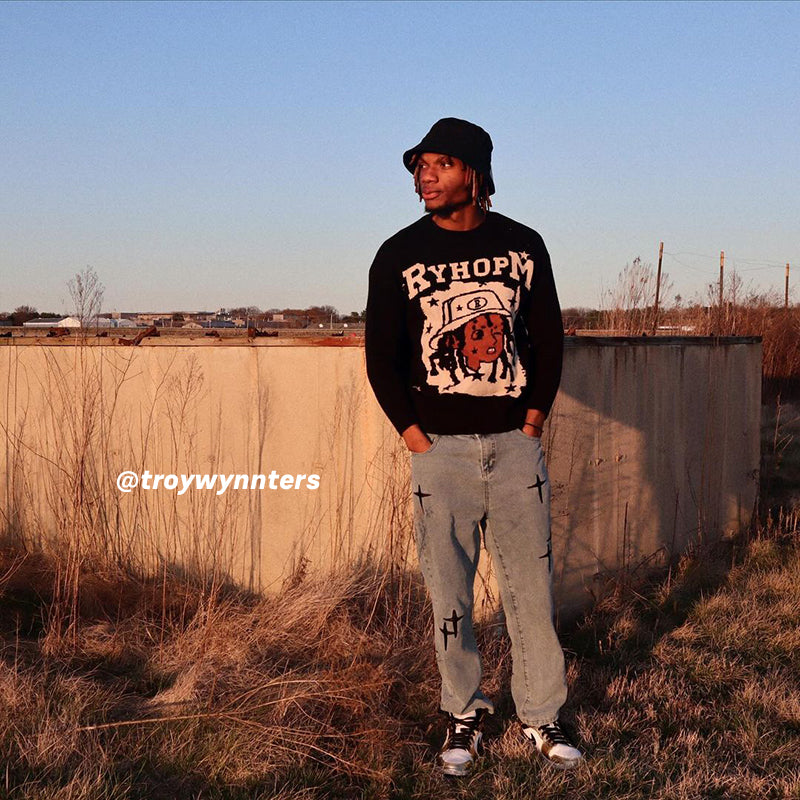 Black RYHOPM graphic sweater, light blue cross-designed jeans, black cap, outdoor field background