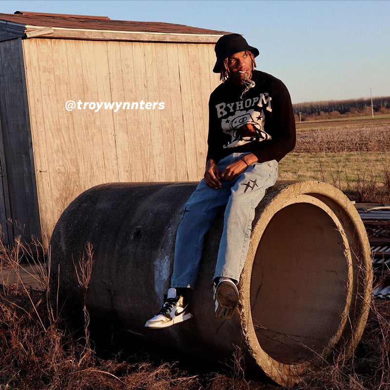 Black RYHOPM graphic sweater, light blue cross-designed jeans, black cap, outdoor field background