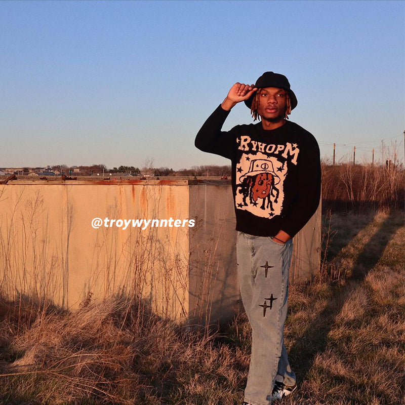 Black RYHOPM graphic sweater, light blue cross-designed jeans, black cap, outdoor field background