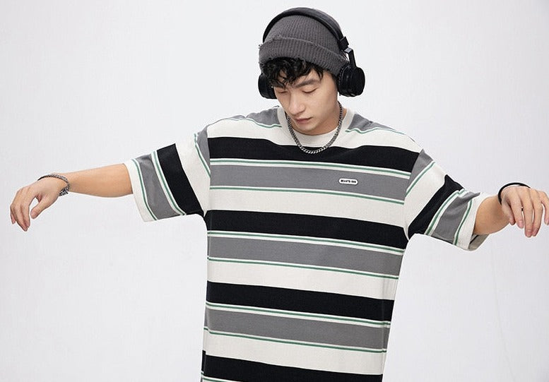 A person wearing a striped shirt with black, gray, and white horizontal lines, a beanie, and headphones stands with arms outstretched against a plain white background. 
