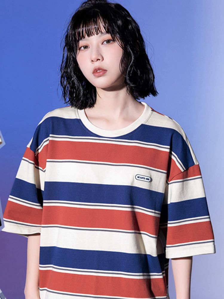 A person with shoulder-length black hair and bangs wearing a red, blue, and beige striped T-shirt with a 'HEART - MO' logo, posing against a solid blue background in a casual streetwear style