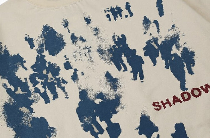 A beige T-shirt with a distressed blue graphic print featuring abstract human figures and the word "SHADOW" in red text on the side, showcasing a bold streetwear design